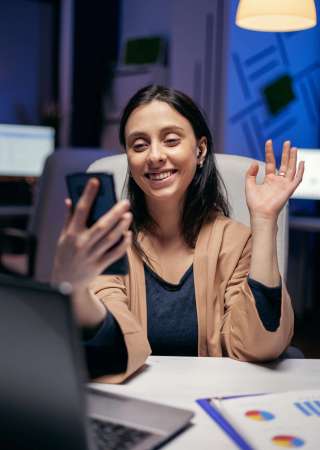 Saying hello in the course of teleconference with entrepreneurs doing overtime. Woman working on finance during a video conference with coworkers at night hours in the office.